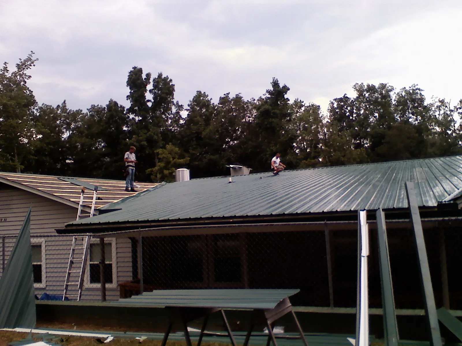 Two roofers installing green metal panels above a covered porch in Andalusia
